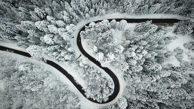Aerial view of a winding road cutting through a dense forest covered in fresh snow in Boege, Auvergne-Rhone-Alpes, France.