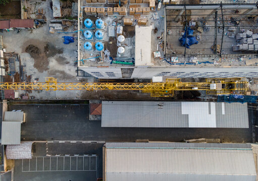 Aerial view of a construction site featuring a yellow tower crane, industrial building with blue water tanks on the roof, and metal-roofed structures in Purwokerto, Central Java, Indonesia.