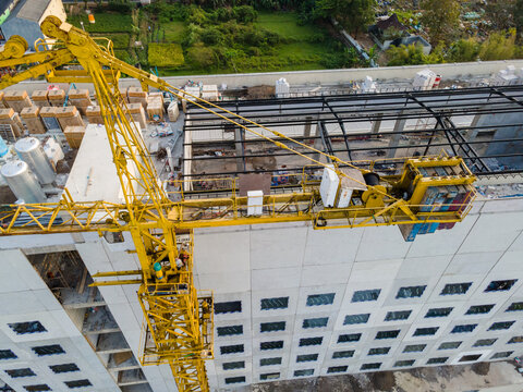 Aerial view of a yellow tower crane at a construction site with concrete building structure and surrounding green fields in Purwokerto, Central Java, Indonesia.