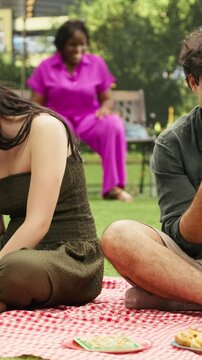Vertical aspect: Boyfriend and girlfriend ignore each other while doomscrolling on smartphones beside a basket, depicting phone dependency, phubbing and disconnection during a picnic date in a park.