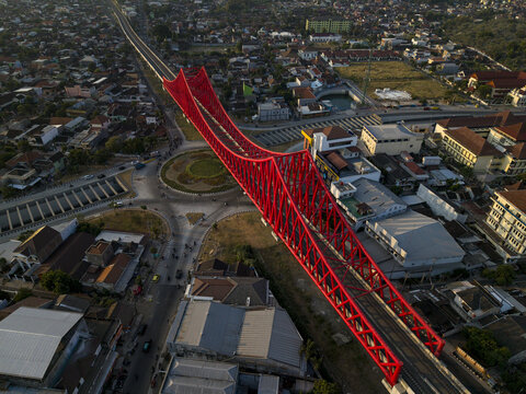 Aerial view of the vibrant red Joglo Railway Bridge spanning over an urban roundabout with surrounding houses and roads in Kecamatan Banjarsari, Jawa Tengah, Indonesia.