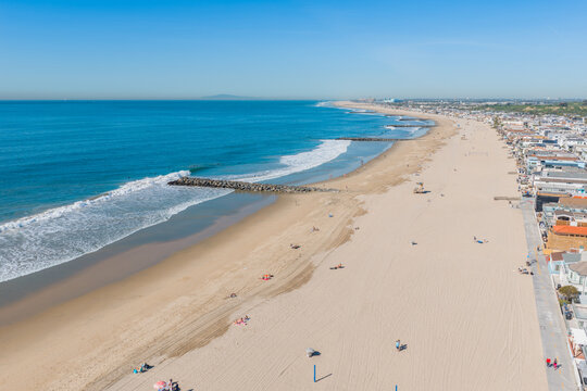 Aerial view of Newport Beach featuring a wide sandy coastline, gentle waves breaking against a stone jetty, and a clear blue sky in Newport Beach, California, United States.