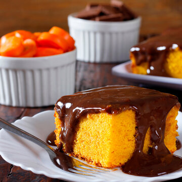 Slice of Brazilian carrot cake topped with creamy chocolate, served on a white plate on a rustic table, with carrots and chocolate in the background.