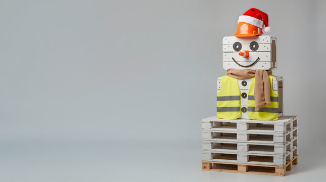 Creative snowman made of wooden pallets and crates wearing a safety vest and hard hat. Industrial Christmas concept with copy space on gray background. Festive warehouse decoration