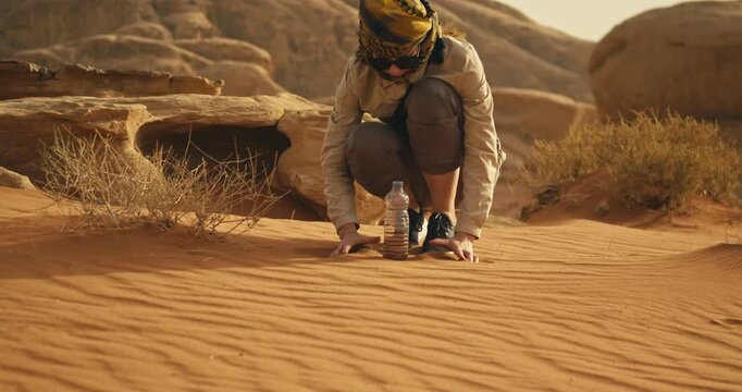 Woman crouching in Wadi Rum, Jordan, carefully adding desert sand into a small container, surrounded by rippled dunes, dry vegetation and rugged sandstone formations.