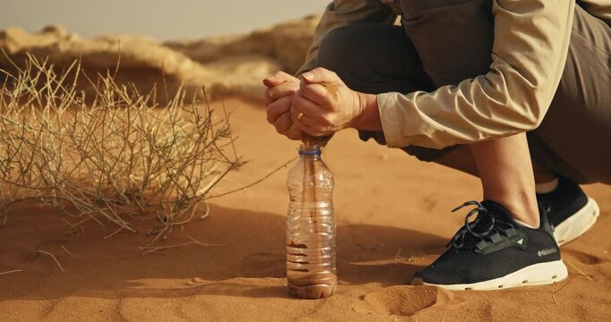 Close up of a person crouching in Wadi Rum, Jordan, carefully filling a clear bottle with desert sand beside dry vegetation in warm natural sunlight.