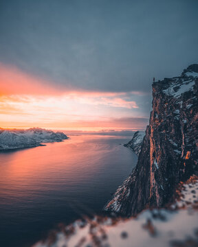 Aerial view of Segla mountain peak with a person standing on the edge overlooking the fjord and snow-covered mountains during a vibrant sunset in Senja, Troms, Norway.