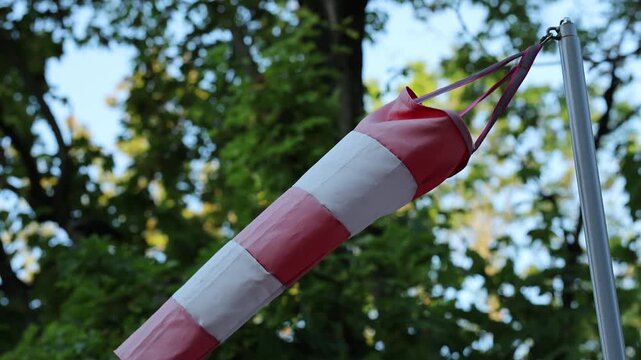 Red and white windsock waving in wind against green trees and blue sky. Meteorological wind cone indicating wind direction and speed. Weather forecast concept.