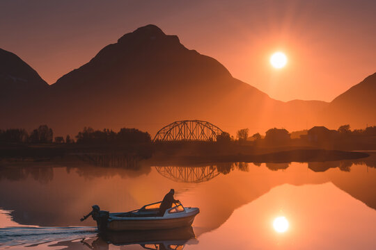 Aerial view of a lone fisherman in a small boat on calm water at sunset, with a bridge and mountain silhouettes in the background in Rodsand, Troms, Norway.