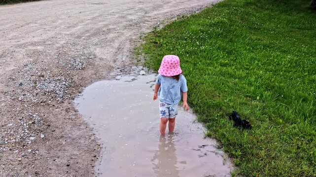 Young child with a cute pink hat jumping and splashing barefoot in a dirty mud puddle