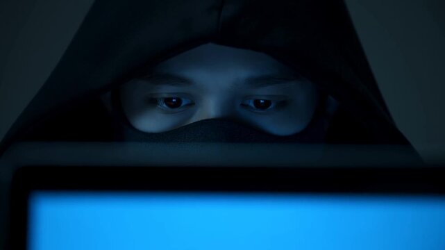 Person in hood and face covering looking at glowing computer screen in dark room