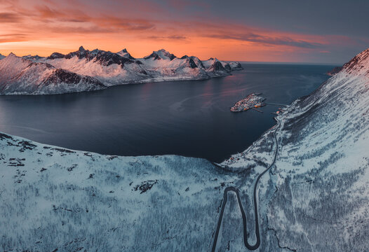Aerial view of the small island village of Husoy nestled in a fjord surrounded by snow-covered mountains and a winding coastal road at sunset in Husoy, Vestfold, Norway.