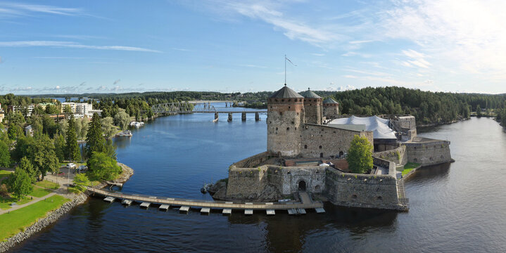 Savonlinna, Finland - 14 August 2026: Aerial view of Olavinlinna Castle surrounded by water, lush green forests, and a bridge in the background under a clear blue sky.