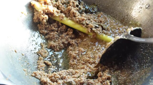 Close-up of hand stirring brown minced meat mixture with spoon in metal wok. Cooking process with oil and spices visible. Authentic culinary scene for food preparation.