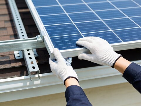 A worker wearing white protective gloves installs a solar panel on a residential roof surface.