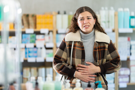 Young woman buyer with stomach pain choosing medicines and hygiene products in pharmacy