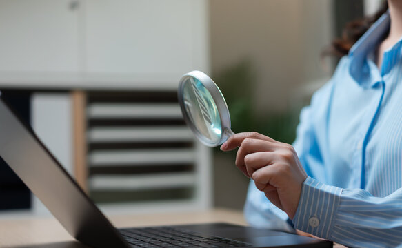 Businesswoman using magnifying glass to inspect laptop data, representing business research, financial analysis, risk assessment, auditing and strategic decision making.