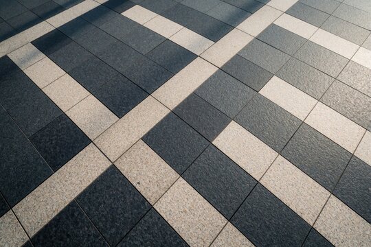 Tiled ground pattern with dark gray and beige stone squares and rectangles. Diagonal light and shadow stripes create geometric shapes on pedestrian walkway surface.