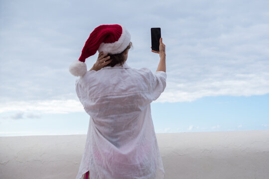 Back view of a woman wearing a red Santa hat and a white shirt on a holiday beach, holding a mobile phone in front of a cloudy sky.