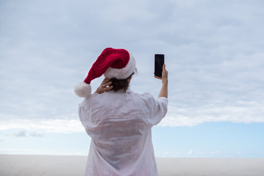 Back view of a woman on holiday wearing a white shirt and a red Santa hat, looking at or taking a picture with a smartphone, with a cloudy sky in the distance