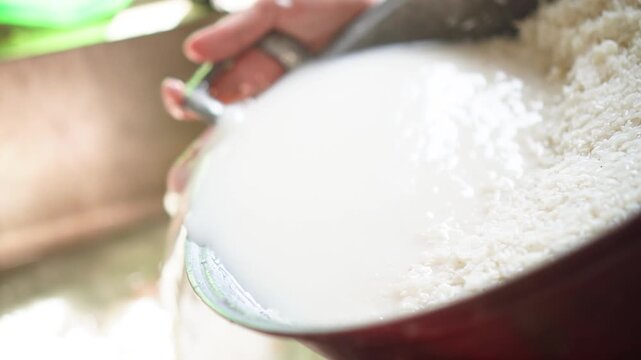 Close-up of hands pouring rice water from a large pot into a bowl, capturing the separation process with natural light and rustic kitchen setting, elegantly showcasing cooking.