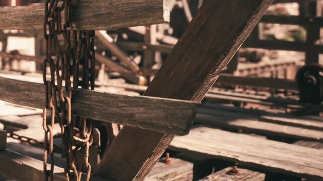 Wooden beams and chains are seen in a warehouse. The scene shows a close up of the structure and its worn materials. Light falls on the wood and metal, highlighting their textures.