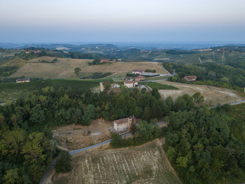 Monforte d'Alba, Piedmont, Italy - 17 August 2026: Aerial view of rolling vineyards and historic buildings in the countryside, featuring lush trees and a quiet road under a clear sky.