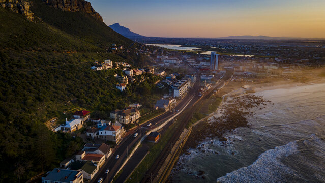 Cape Town, Western Cape, South Africa - 17 August 2026: Aerial view of Muizenberg at sunset, showing the coastal road, railway, hillside houses, and waves hitting the rocky shore.