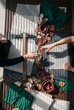 Overhead view of designers reaching towards each other over a wooden desk with ceramic shards, drafting tools, and tracing paper under sunlight shadows