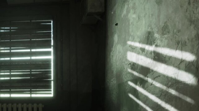 Sunlight streams through closed shutters, casting shadows on the wall in a room that has worn paint. The atmosphere shows signs of age and neglect.