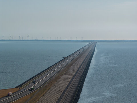 Aerial view of the Afsluitdijk causeway stretching across the water with a highway, traffic, and wind turbines in the distance in Breezanddijk, Friesland, Netherlands.