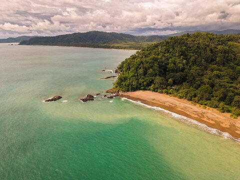 Aerial view of the lush tropical rainforest meeting the turquoise ocean waters, with rocky outcrops along the sandy coastline under a cloudy sky in Drake Bay, Costa Rica.
