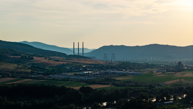 Aerial view of the Mintia power plant with its distinctive chimneys and cooling towers set against rolling hills and agricultural fields in Deva, Hunedoara, Romania.