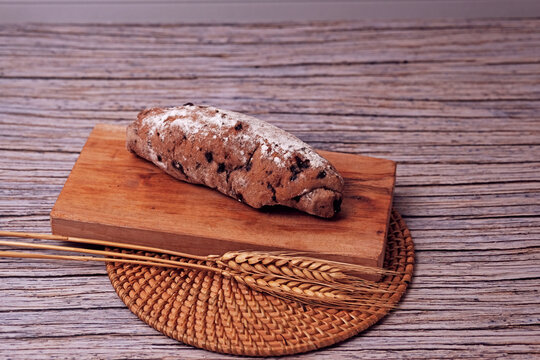 Fresh sourdough rests on wooden board, dusted with flour. Warm bread sits atop rustic cutting board. Wheat stalks lie beside it, woven mat below. Subtle shadow hints at soft, cozy kitchen light