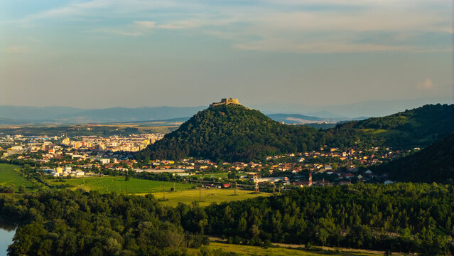 Aerial view of Deva Fortress perched on a hill overlooking the town with surrounding green fields, forests, and residential areas in Deva, Hunedoara, Romania.