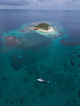 Aerial view of a small tropical island with palm trees and buildings surrounded by clear turquoise water with a sailboat and dinghy anchored nearby in Guna Yala Comarca, Panama.