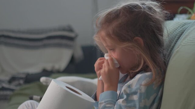 A young girl sits in her living room, feeling sick and holding a tissue. She looks around, seeking warmth and care while wrapped in a blanket on a soft couch.