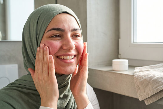 Smiling hijab woman applying moisturizer in bright bathroom skincare routine