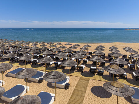 Aerial view of Marina de Vilamoura beach with rows of straw parasols and sun loungers on golden sand overlooking the Atlantic Ocean in Quarteira, Faro, Portugal.