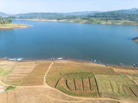 Aerial view of Wadaslintang reservoir with small fishing boats moored along the shore, surrounded by agricultural fields and rolling hills in Wadaslintang, Central Java, Indonesia.