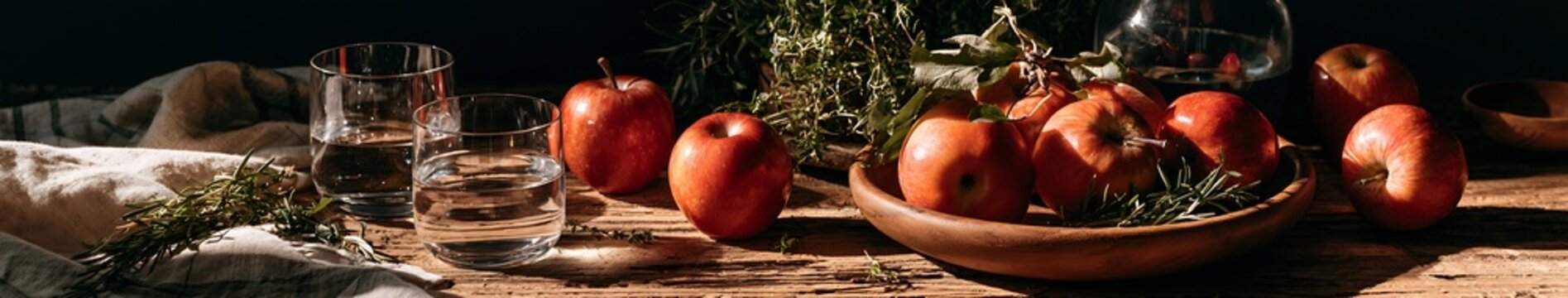 Red apples and fresh herbs on a rustic wooden table with glass tumblers and linen &mdash; autumn still life banner