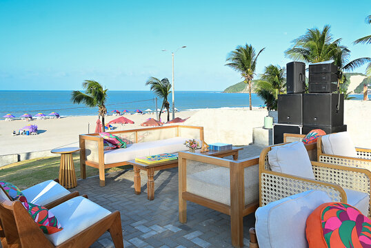 outdoor beach lounge deck with wooden armchairs and colorful patterned cushions, seaside event setup on paved stone terrace with ocean view and palm trees