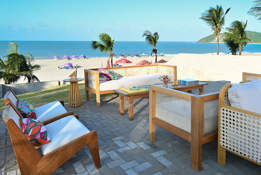 outdoor beach lounge deck with wooden armchairs and colorful patterned cushions, seaside event setup on paved stone terrace with ocean view and palm trees