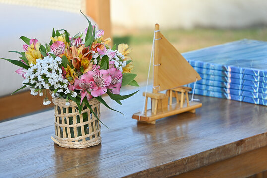 yellow flower bouquet in woven glass vase and miniature wooden sailboat models on coffee table, rustic beach event setup with wicker lounge chair in background