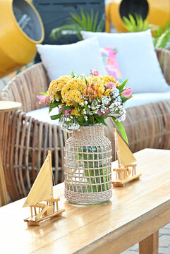 yellow flower bouquet in woven glass vase and miniature wooden sailboat models on coffee table, rustic beach event setup with wicker lounge chair in background