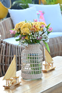yellow flower bouquet in woven glass vase and miniature wooden sailboat models on coffee table, rustic beach event setup with wicker lounge chair in background