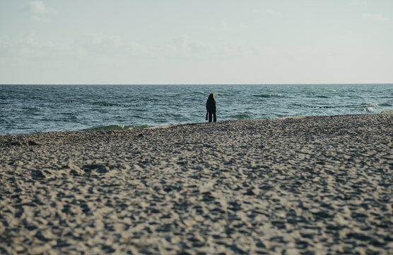 Woman standing and walking on empty sandy beach. Person alone at seashore with waves. Solitude, freedom and relaxation by the sea. Autumn vacation concept