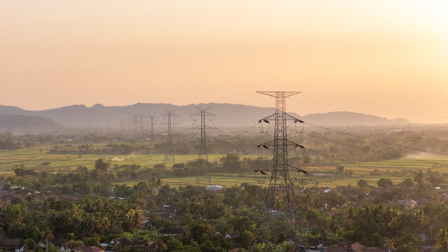 Aerial view of high-voltage power transmission towers stretching across lush green rice fields and rural village houses in the golden light of sunset in Klaten, Central Java, Indonesia.