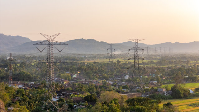 Aerial view of electricity pylons standing over a rural landscape with houses, lush trees, and distant mountains under a warm golden hour sky in Klaten, Central Java, Indonesia.