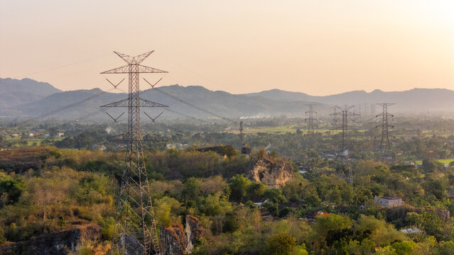 Aerial view of high-voltage electricity pylons standing over a rural landscape with lush trees, rocky outcrops, and distant hills in Klaten, Central Java, Indonesia.